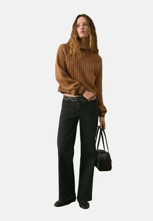 Maglione marrone a maglia con texture a coste, abbinato a jeans neri con cintura decorata da borchie. Il modello tiene in mano una borsa nera.