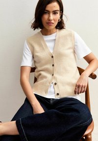 Gilet a maglia beige con scollo a V e bottoni neri, indossato sopra una T-shirt bianca. Il soggetto è seduto contro un muro chiaro su una sedia di legno.