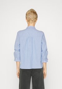 Una persona con capelli corti indossa una camicia a righe azzurre con maniche arruffate, girata di spalle su uno sfondo bianco uniforme.