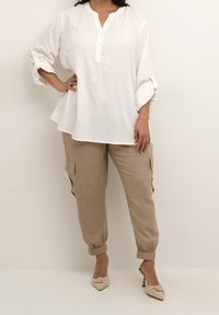 Chemise blanche en lin à boutons avec col Henley, associée à un pantalon cargo beige avec poches latérales et poignets élastiques. Des chaussures à talons beiges complètent le look.