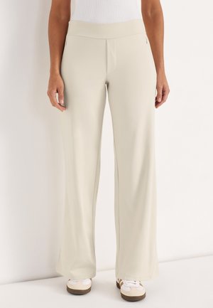 Frau trägt cremefarbene Hosen mit weitem Bein und beigen Turnschuhen, steht vor einem einfarbigen hellen Hintergrund.