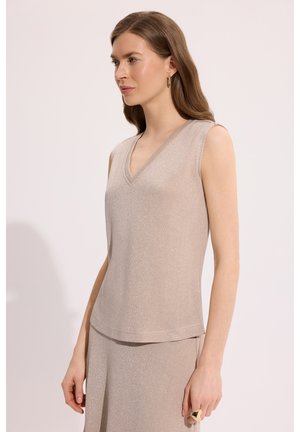 Kobieta z długimi brązowymi włosami nosi beżowy, bezrękawnik z dekoltem w serek i pasującą spódnicę, uzupełnione złotymi okrągłymi kolczykami i pierścionkiem.