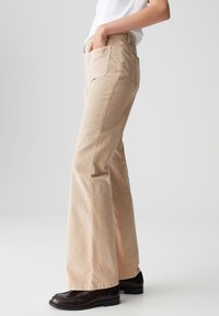 Persona che indossa pantaloni beige a gamba larga e mocassini marrone scuro, in piedi con una mano in tasca contro uno sfondo bianco semplice.