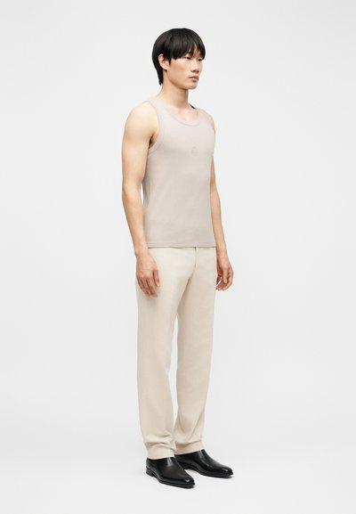 Άνδρας στέκεται πλαγίως φορώντας αμάνικο τοπ μπεζ, ανοιχτό μπεζ παντελόνι και μαύρα δερμάτινα παπούτσια, μπροστά σε απλό ανοιχτό φόντο.