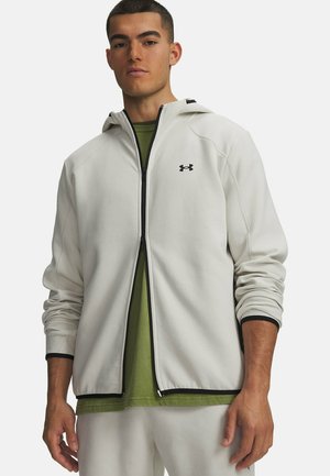 Jonge man draagt een lichtgrijze zip-up hoodie over een olijfgroen shirt, staand met de handen ontspannen langs zijn zij.