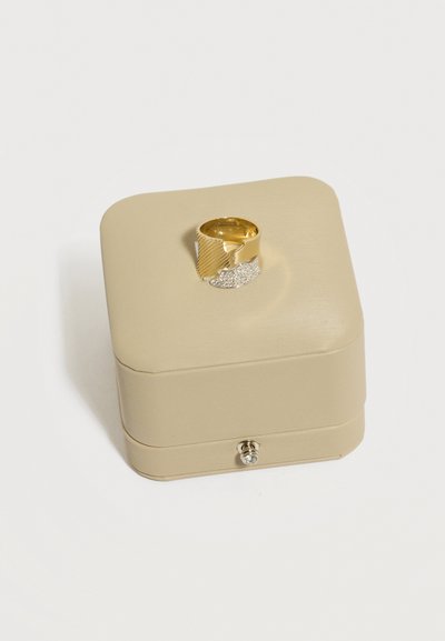 Bague à large anneau texturée en or et argent présentée sur une boîte à bijoux carrée beige avec un petit fermoir en pierre précieuse.
