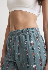 Ljusa teal-pyjamasbyxor med ett festligt nötknäppartema. Har resårliv och en mjuk textur. Mönstret inkluderar olika nötknäppardesigner.
