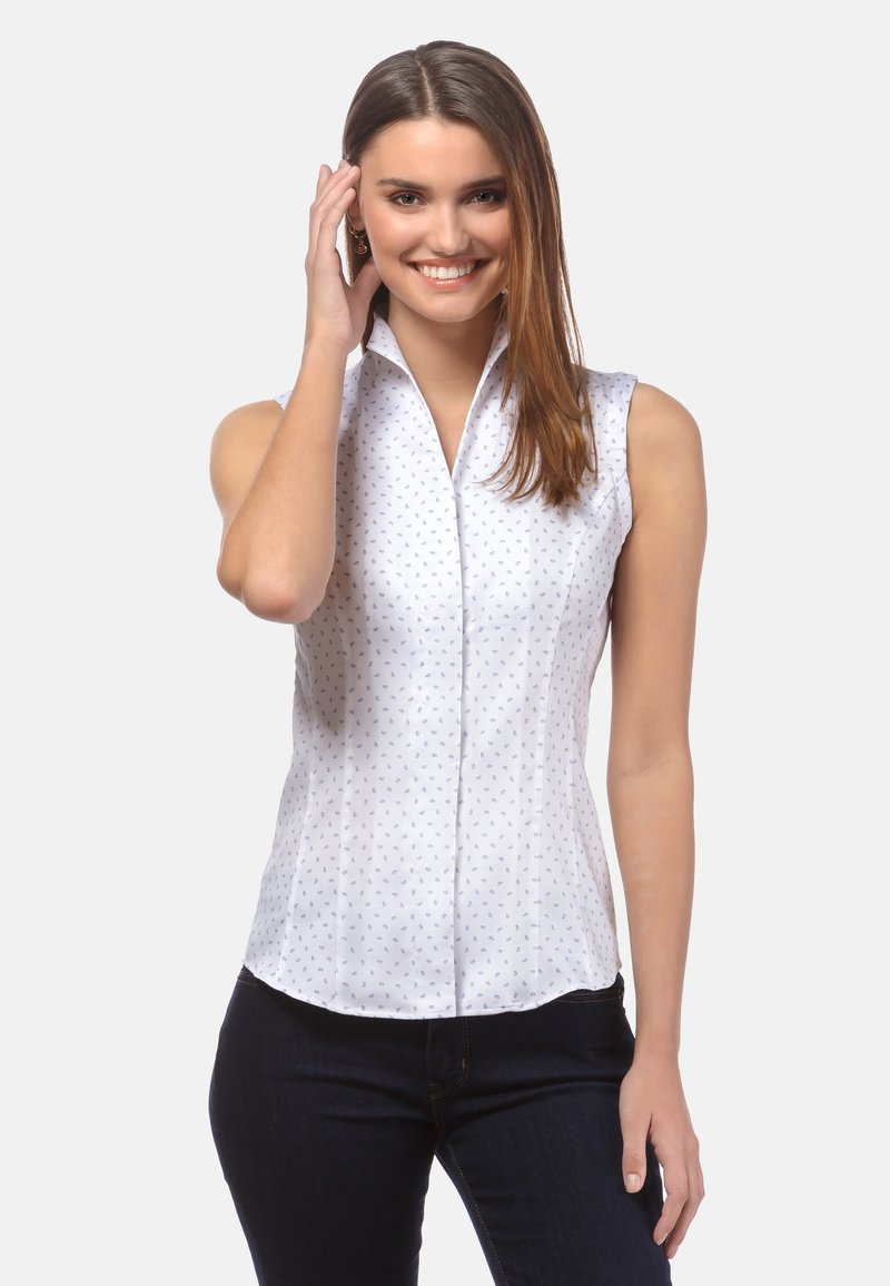 Blusa bianca senza maniche con un delicato motivo floreale blu, colletto alto e chiusura con bottoni; realizzata in tessuto leggero. Indossata con jeans scuri.