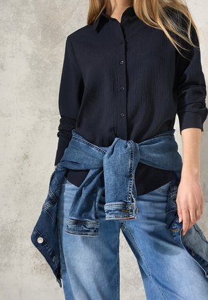 Femme portant une chemise noire à manches longues boutonnée avec une veste en jean nouée autour de la taille et un jean bleu clair.