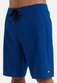 Blå badshorts tillverkade av lättviktig tyg, med en svart dragsko, en patch-logotyp på nedre vänstra delen, och en slät yta.