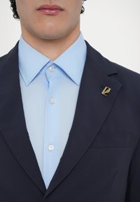 Navy blézer világoskék inggel. Az ing gombos gallérral és ezüstgombbal rendelkezik, arany hajtókával díszítve.