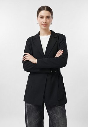 Kvinne står med armene krysset, iført en svart blazer over en hvit topp og mørkegrå jeans mot en ensfarget lys bakgrunn.