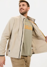 Beige fleecejakke med sort lynlås, dekoreret med sidelommer og elastiske manchetter. Båret over en lysegrøn t-shirt med orange tekst.
