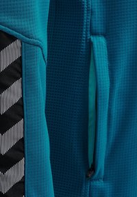 Chaqueta deportiva en color verde azulado con tejido texturizado, detalle a rayas en negro y blanco, y un bolsillo con cremallera en el lateral para almacenamiento.