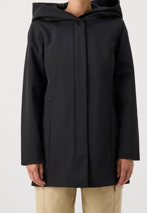 Osoba ubrana w czarny płaszcz z kapturem z ukrytym zapięciem z przodu i zapinanymi na zamek kieszeniami po bokach, w połączeniu z beżowymi spodniami na jednolitym tle.