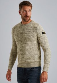 Pull en tricot beige clair avec un motif texturé, col rond et poignets côtelés ; présente une petite étiquette noire sur la manche.