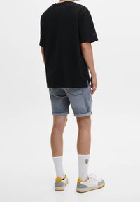 Persona que lleva una camiseta negra, pantalones cortos de mezclilla gris remangados, calcetines blancos con un diseño de palmera y zapatillas blancas con detalles amarillos, de espaldas.