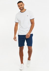 Camiseta blanca de manga corta con acentos en azul marino, combinada con pantalones cortos de mezclilla azul oscuro. El modelo lleva zapatillas blancas y está de pie con una sonrisa.