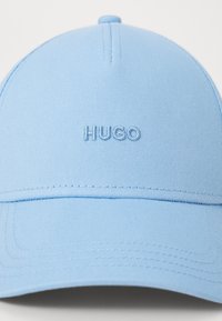 Lys blå baseballcap lavet af stof, med broderet "HUGO" logo på fronten, med en buet skygge og syet detaljer.