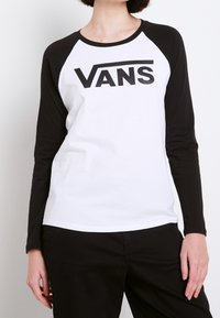 Bílá a černá dlouhá rukávová raglánová trička s velkým černým logem "VANS" přes hrudník. Vyrobeno z měkké bavlněné tkaniny.