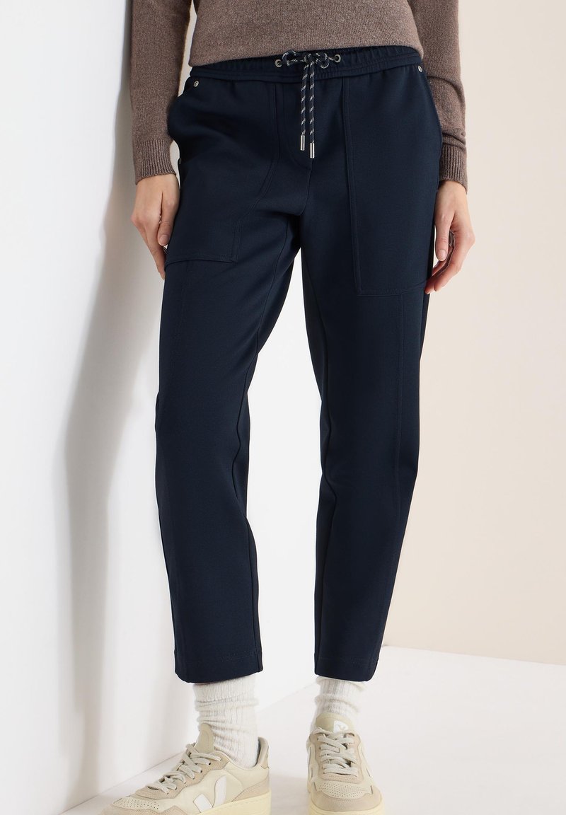 Pantalon de jogging bleu marine avec une taille élastique, des poches avant et une coupe décontractée. Porté avec des baskets beiges et des chaussettes blanches.