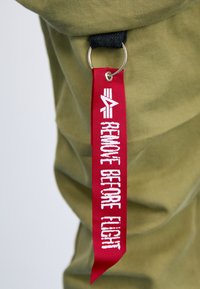 Groene stoffen tas met een zwarte schouderriem en een rood stoffen label dat in witte letters "VERWIJDER VOORDAT JE VLIEGT" leest, bevestigd met een metalen ring.