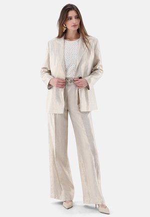 Donna che indossa un completo crema testurizzato con pantaloni a gamba larga, cintura, top bianco con fantasia, orecchini dorati vistosi e tacchi appuntiti crema.