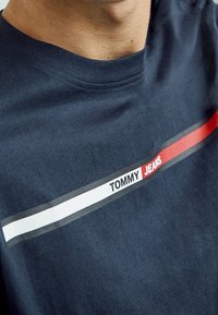 Námořnické modré bavlněné tričko s kulatým výstřihem, s vodorovným designem červených, bílých a šedých pruhů a textem "TOMMY JEANS" v bílé barvě.