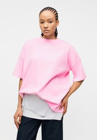 Νεαρό άτομο με κοτσίδες cornrow που φοράει υπερμεγέθη ροζ μπλούζα πάνω από λευκή μπλούζα και σκούρο παντελόνι, ποζάροντας με το χέρι στη μέση μπροστά σε λευκό φόντο.