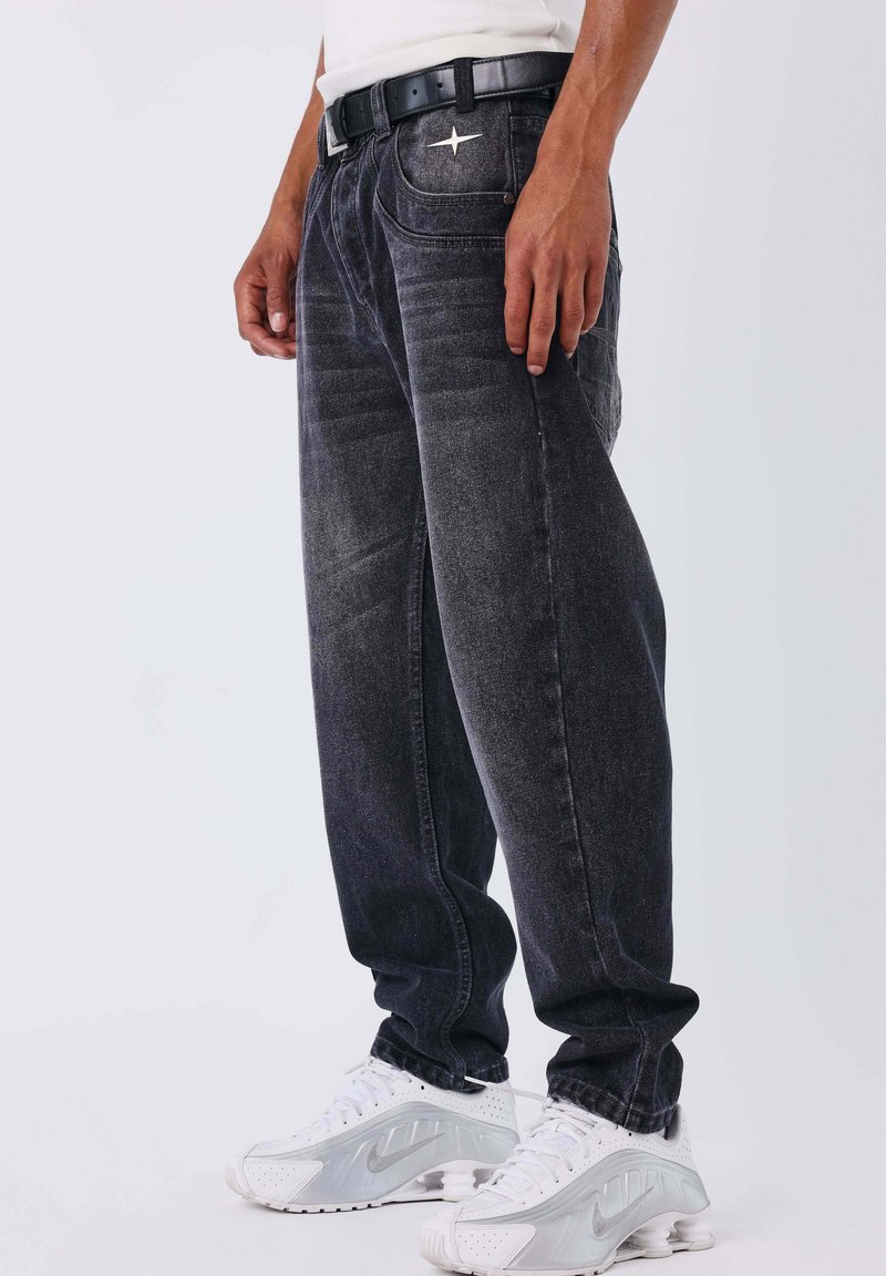 Jeans in denim grigio scuro con vestibilità a gamba dritta, caratterizzati da un accento a stella argentata sul davanti. Indossati con una cintura nera e scarpe da ginnastica bianche.