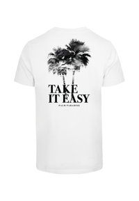 White cotton t-shirt featuring a graphic of palm trees and the text "TAKE IT EASY PALM PARADISE" in bold black lettering.