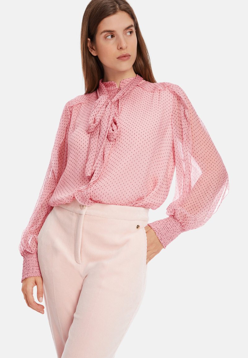 D blouse mit rosa Punkten, einem hohen Kragen, einer Schleifen-Detail und voluminösen Ärmeln, kombiniert mit hellrosa Cordhosen.