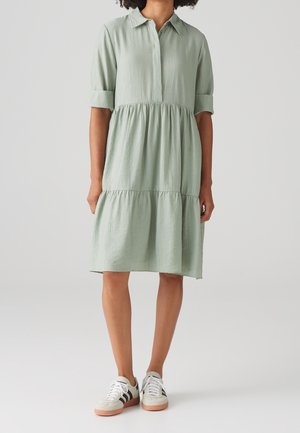 Femme portant une robe verte claire à plusieurs volants, longueur genou, avec des manches retroussées aux trois quarts et des baskets blanches à rayures noires.