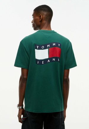 Uomo che indossa una t-shirt verde Tommy Jeans con il logo sulla schiena, in piedi davanti a uno sfondo chiaro e uniforme.
