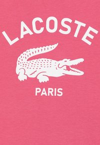 Ružičasta pamučna majica s bijelim natpisom "LACOSTE" i grafikom krokodila, sa natpisom "PARIS" ispod. Glatka tekstura, ležeran dizajn.