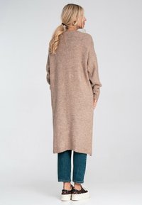 Kobieta z blond włosami w kitce, ubrana w długi beżowy sweter, niebieskie dżinsy i trampki w panterkę, stojąca na tle czystego białego tła.