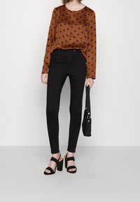 Femme portant un chemisier marron à manches longues avec des pois noirs, un pantalon skinny noir, des sandales à talons hauts noires, tenant un sac à main noir.