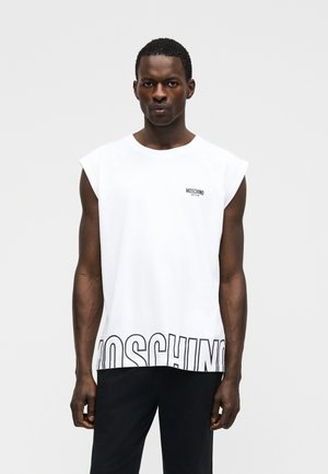Mężczyzna ubrany w białą, bezrękawnikową koszulkę Moschino z czarnym napisem na wysokości klatki piersiowej i dużym konturowym napisem na dole, w połączeniu z czarnymi spodniami.