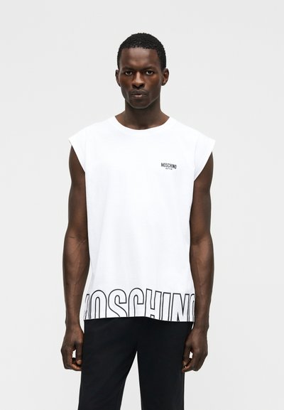 Άνδρας που φοράει λευκό αμάνικο μπλουζάκι Moschino με μαύρο λογότυπο κειμένου στο στήθος και μεγάλο περιγράμμα κειμένου στο τελείωμα, συνδυασμένο με μαύρο παντελόνι.
