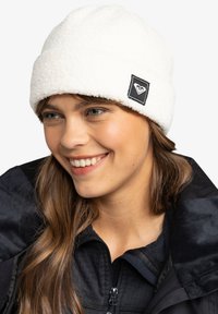 Witte fleece beanie met een omgeslagen boord en een zwart vierkant logo patch. Zachte textuur en ronde vorm, geplaatst op het hoofd van een persoon.