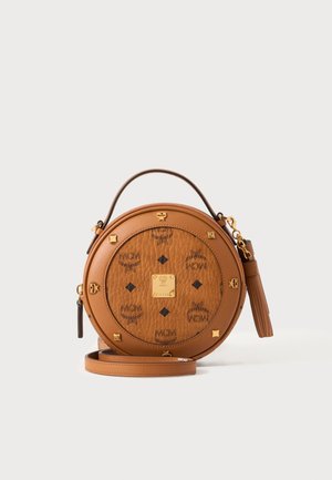 Sac à main rond en cuir marron avec clous dorés, impression du logo MCM, poignée supérieure, tirette de fermeture éclair à pompon et bandoulière amovible sur fond blanc.