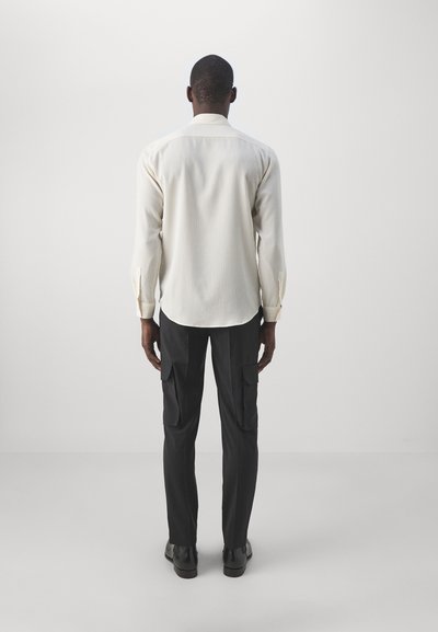 Man staat met zijn rug naar de camera, gekleed in een crèmekleurig shirt met lange mouwen, zwarte cargobroeken en zwarte schoenen, tegen een effen grijze achtergrond.