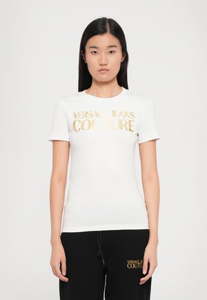 T-shirt blanc à manches courtes avec un col rond, arborant le logo en feuille d'or "VERSACE JEANS COUTURE" sur la poitrine. Design lisse et ajusté.
