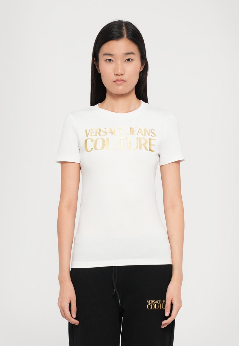 Camiseta blanca de manga corta con cuello redondo que presenta el logo dorado en foil de "VERSACE JEANS COUTURE" en el pecho. Diseño suave y ajustado.