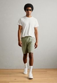 Witte t-shirt, groene shorts en witte sneakers. De outfit heeft een ontspannen pasvorm met een elastische tailleband en wordt gedragen met een zonnebril.