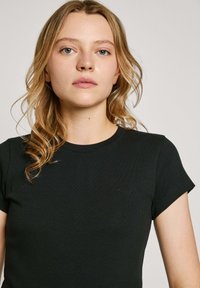 Blonde Frau mit welligem Haar trägt ein eng anliegendes, schwarzes, kurzärmeliges, geripptes Shirt und steht vor einem schlichten hellen Hintergrund.