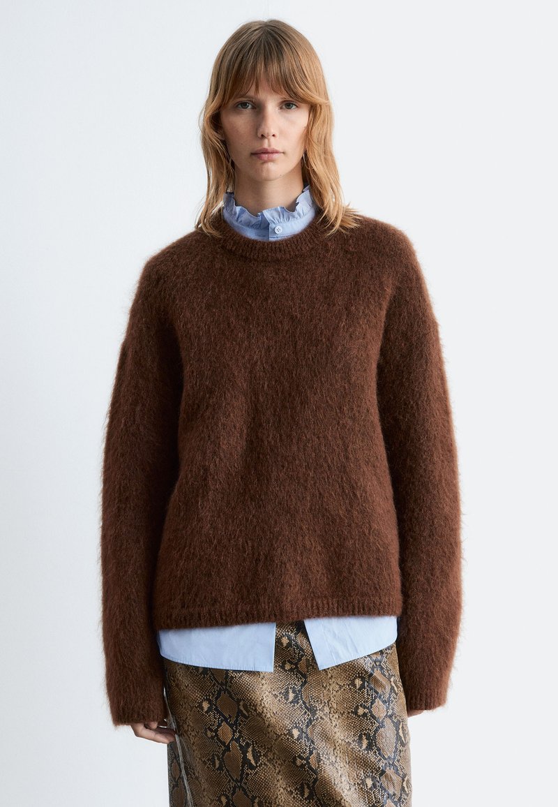 Brauner, flauschiger Pullover mit rundem Halsausschnitt und langen Ärmeln, getragen über einem hellblauen Hemd und einem braunen, schlangenbeschichteten Rock.