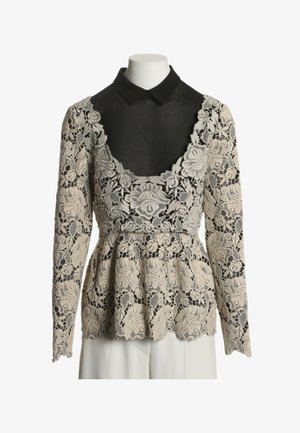 Haut en dentelle florale beige à manches longues sur chemise noire à col, taille ajustée, ourlet festonné, présenté sur un torse de mannequin blanc.