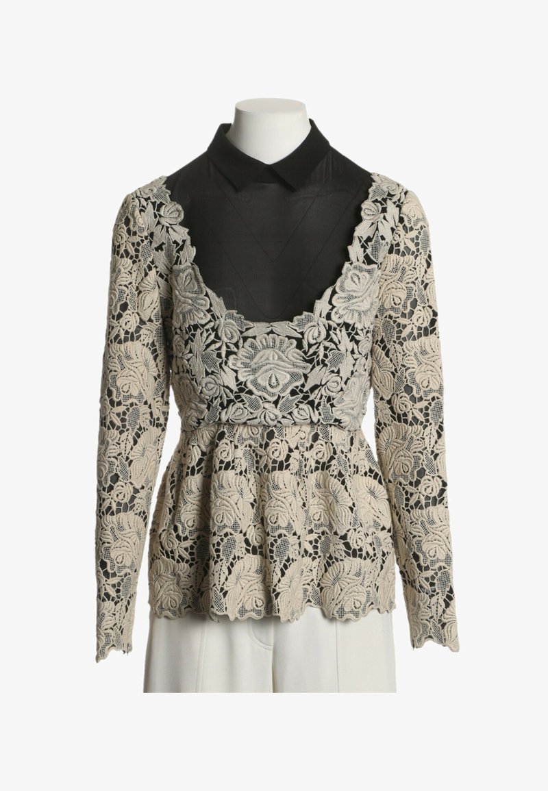 Haut en dentelle florale beige à manches longues sur chemise noire à col, taille ajustée, ourlet festonné, présenté sur un torse de mannequin blanc.