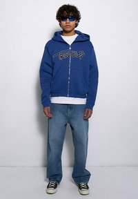 Niebieska bluza z zamkiem błyskawicznym z grafiką "TROUBLE", zestawiona z luźnymi niebieskimi dżinsami i czarnymi sneakersami. Model nosi okulary przeciwsłoneczne.